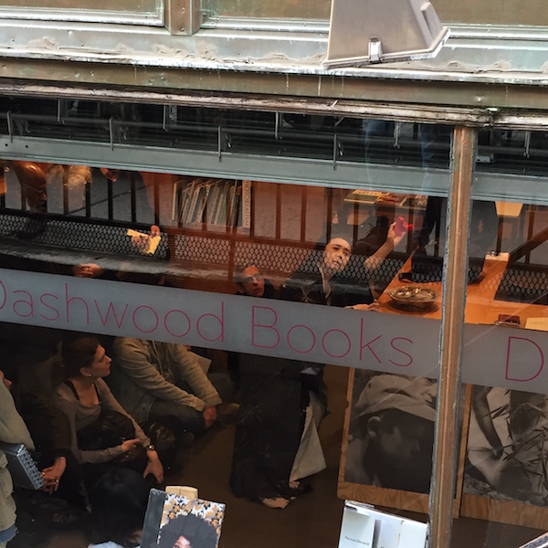 We see Eiko perform in the bookstore through the store's front window. Her hands are raised.